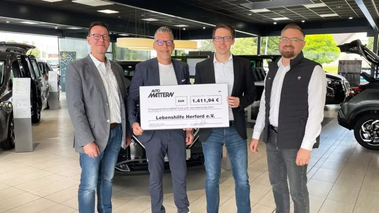 Vier Männer stehen in einem Autohaus bei Auto Mattern und halten einen großen Spende-Scheck über 1.411,94 Euro, ausgestellt auf die Lebenshilfe Herford e.V. Im Hintergrund sind Autos zu sehen.