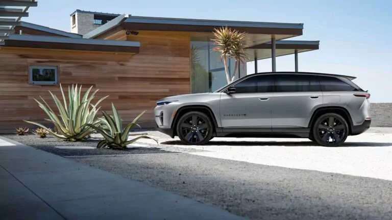 Ein silberner Jeep Wagoneer Elektro SUV ist vor einem modernen Holzhaus mit großen Glasfenstern geparkt. Wüstenpflanzen und Schotterflächen umgeben die Einfahrt unter einem klaren Himmel. Informieren Sie sich über die neuesten Elektro SUV-Nachrichten.