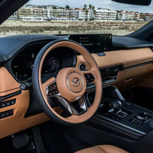 Innenansicht eines modernen Mazda-Fahrzeugs mit hellbraunen Ledersitzen, einem schwarzen Lenkrad mit dem Mazda-Logo und einem digitalen Display – all dies präsentiert im innovativen Mazda CX-80 als Plug-in-Hybrid – entdecken Sie die Möglichkeiten. Genießen Sie durch die Windschutzscheibe den Blick auf Gebäude und Palmen, die dieses stilvolle und umweltfreundliche Fahrerlebnis perfekt ergänzen.