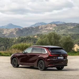 Ein kastanienbrauner Mazda CX-80 als Plug-in-Hybrid steht auf einer gepflasterten Fläche, im Hintergrund sind grüne Hügel, Bäume und Berge zu sehen, über ihnen ein bewölkter Himmel – entdecken Sie die Möglichkeiten.