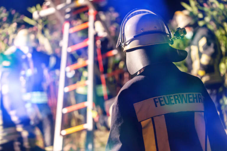 Ein Feuerwehrmann in Schutzkleidung mit der Aufschrift Feuerwehr steht bei einem nächtlichen Einsatz in der Nähe einer Leiter mit verschwommenen Kollegen und Notbeleuchtung im Hintergrund.