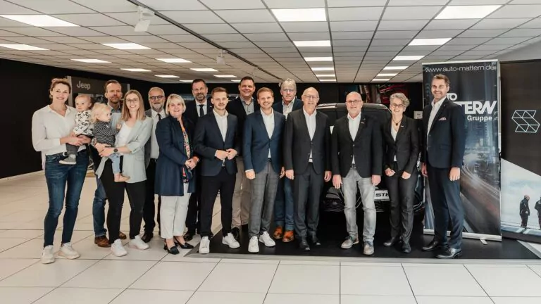 Eine Gruppe von sechzehn Erwachsenen, einige in Geschäftskleidung und einer mit einem Kind im Arm, lächeln für ein Gruppenfoto in einem modernen, gut beleuchteten Ausstellungsraum von Auto Mattern mit Preisauszeichnungsbannern und Autos im Hintergrund.