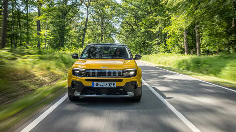 Ein gelber Jeep Avenger 4xe SUV fährt an einem sonnigen Tag eine von Bäumen gesäumte Straße hinunter, umgeben von üppigem grünen Laub. Das Foto ist aus der Frontperspektive aufgenommen und zeigt den Kühlergrill und die Scheinwerfer des Fahrzeugs.