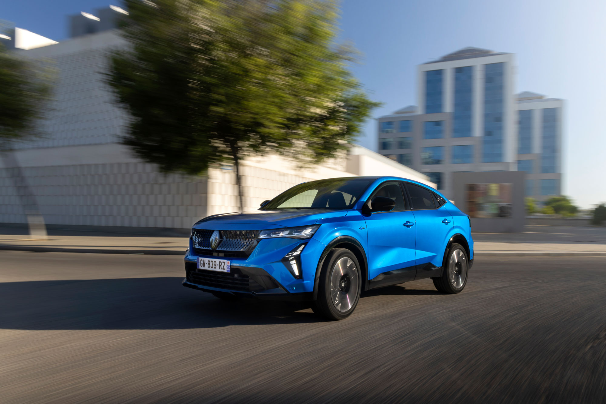 Ein hellblauer Renault Rafale SUV fährt auf einer Stadtstraße mit verschwommenen Bäumen und modernen weißen Gebäuden im Hintergrund, was auf Bewegung und eine städtische Umgebung hindeutet - perfekt für Ihr nächstes Gewerbe Angebot Leasing.