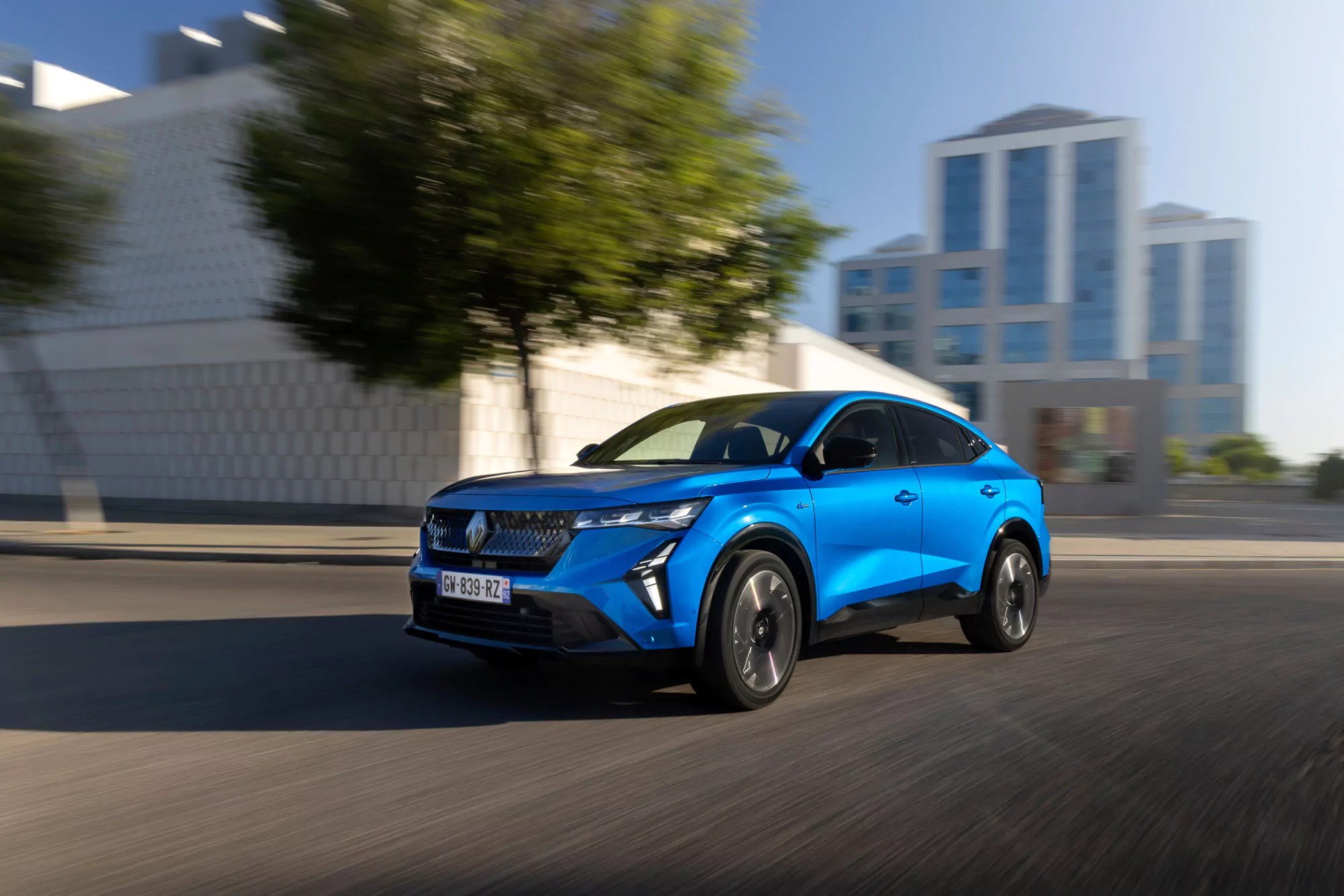 Ein hellblauer Renault Rafale SUV fährt auf einer Stadtstraße mit verschwommenen Bäumen und modernen weißen Gebäuden im Hintergrund, was auf Bewegung und eine städtische Umgebung hindeutet - perfekt für Ihr nächstes Gewerbe Angebot Leasing.