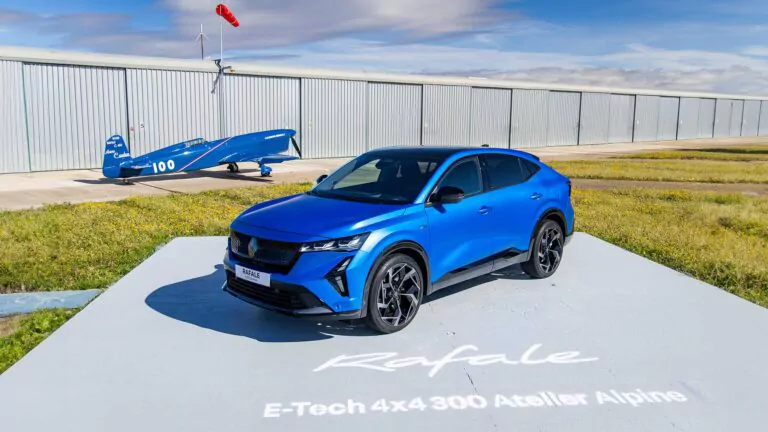 Ein blauer Renault Rafale E-Tech Plug-in-Hybrid-Geländewagen ist auf einer Plattform mit der Aufschrift Rafale E-Tech 4x4 300 Atelier Alpine geparkt. Im Hintergrund steht ein blaues Oldtimer-Flugzeug in der Nähe eines großen Hangars unter einem teilweise bewölkten Himmel.