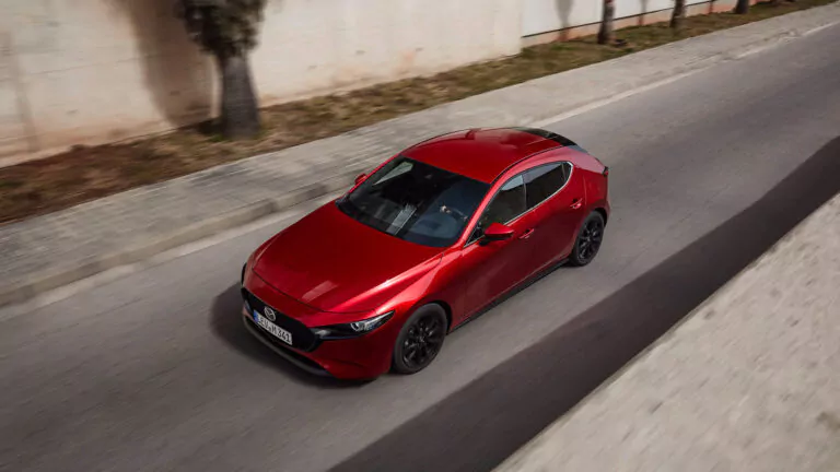 Ein glänzender roter Schrägheckwagen fährt auf einer gepflasterten Straße neben einem Gehweg mit Bäumen und einer Betonmauer, von oben gesehen.