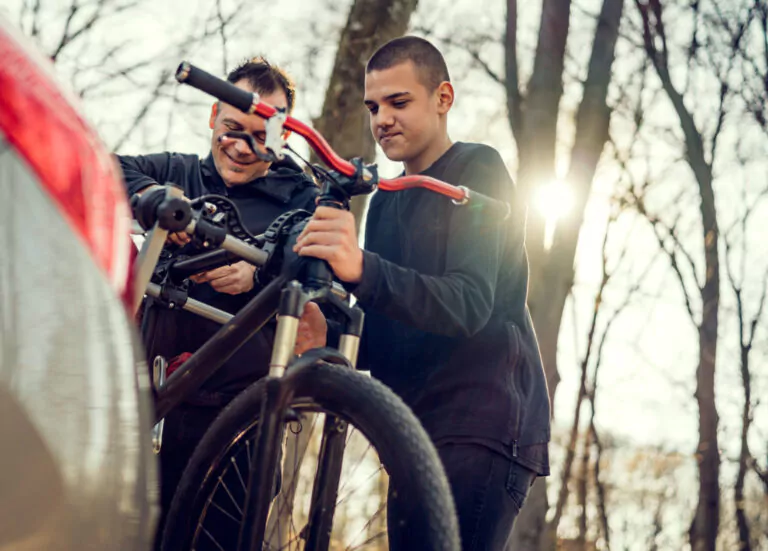 Zwei Personen befestigen ein Fahrrad an einem Autofahrradträger in einem Waldgebiet bei Tageslicht, das Sonnenlicht fällt durch die Bäume - so sind Sie mit Auto Mattern sicher unterwegs: Fahrrad richtig transportieren!.