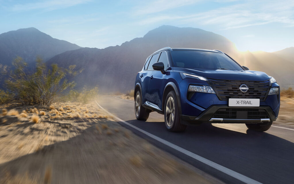 Ein blauer Nissan X-Trail N-Connecta fährt auf einer asphaltierten Straße durch eine Wüstenlandschaft mit Bergen im Hintergrund unter einem klaren Himmel – eine ideale Kulisse, um den Komfort und das Abenteuer eines Nissan X-Trail N-Connecta im Leasing zu erleben.