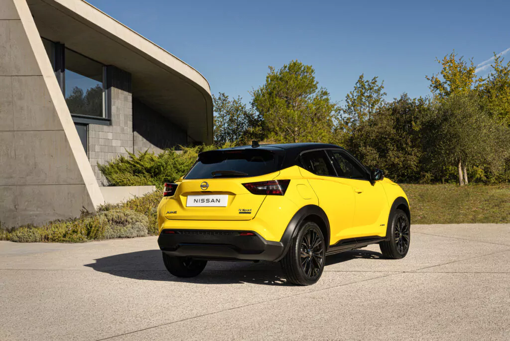 Ein leuchtend gelber und schwarzer Nissan Juke parkt auf einer Einfahrt in der Nähe eines modernen Betongebäudes, umgeben von Grün und Bäumen unter einem klaren blauen Himmel.