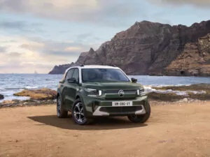 Ein grüner Citroen C3 Aicross Geländewagen, geparkt an einer Sandküste mit Felsen und Klippen im Hintergrund unter einem teilweise bewölkten Himmel.