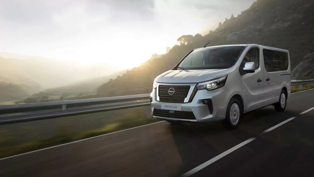 Ein weißer Nissan Primastar Kombi fährt auf einer asphaltierten Bergstraße bei Sonnenaufgang, mit nebligen Hügeln und Sonnenlicht im Hintergrund - entdecken Sie unschlagbare Nissan Primastar Kombi Angebote für Ihr nächstes Abenteuer.