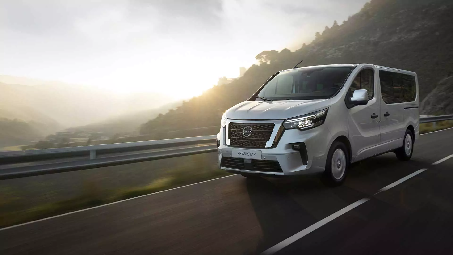 Ein weißer Nissan Primastar Kombi fährt auf einer asphaltierten Bergstraße bei Sonnenaufgang, mit nebligen Hügeln und Sonnenlicht im Hintergrund - entdecken Sie unschlagbare Nissan Primastar Kombi Angebote für Ihr nächstes Abenteuer.