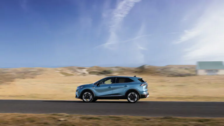 Ein blauer Geländewagen fährt auf einer asphaltierten Straße durch eine weite, offene Landschaft mit trockenem Gras und sanften Hügeln unter einem teilweise bewölkten Himmel.