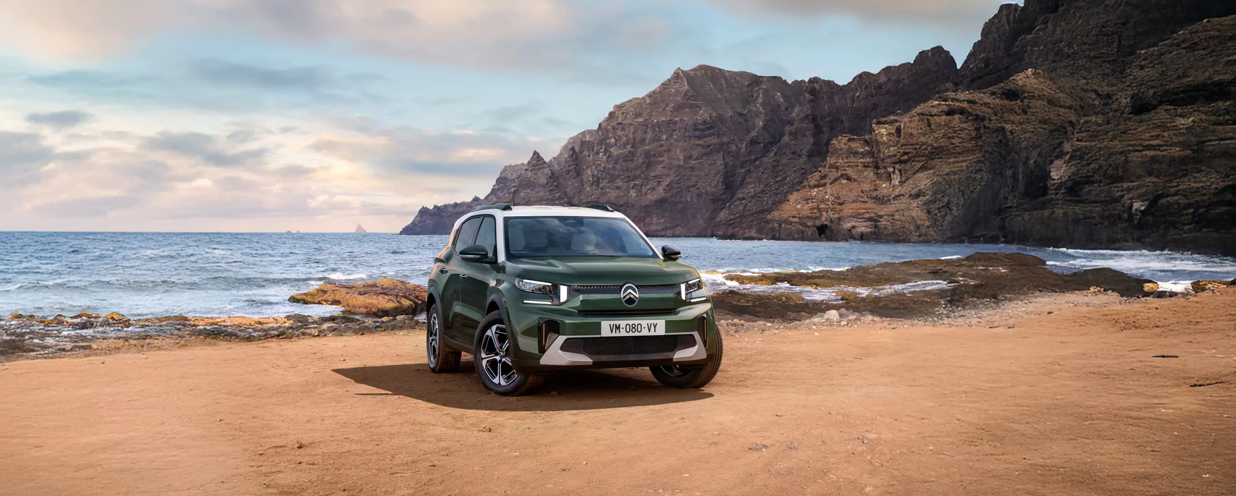 Ein Citroen C3 Aircross, grün mit weißem Dach, steht an einem sandigen, felsigen Ufer mit dramatischen Klippen und dem Meer im Hintergrund unter einem teilweise bewölkten Himmel.