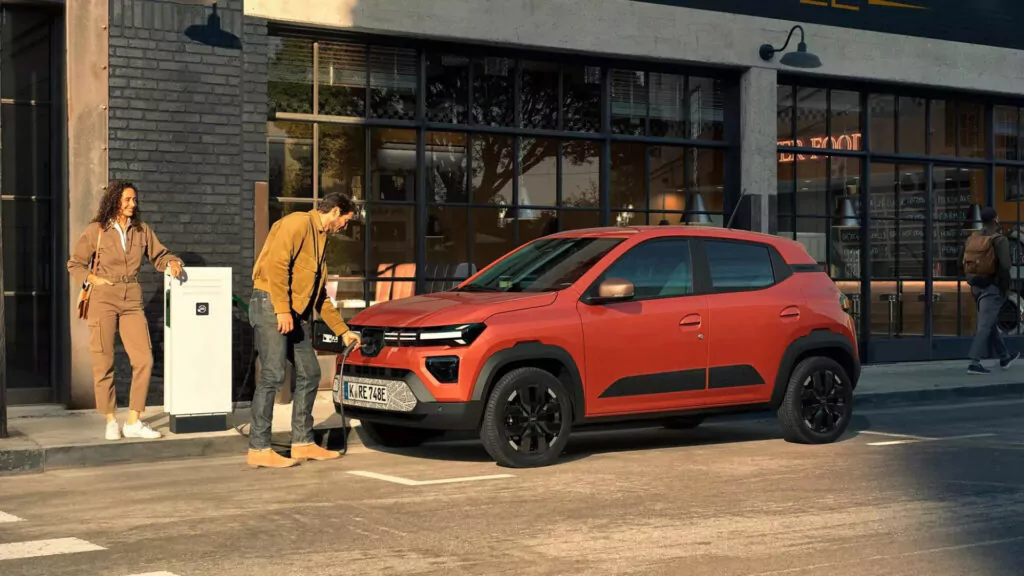 Ein Mann schließt ein orangefarbenes Elektroauto an eine Ladestation in einer Stadtstraße an, während eine Frau lächelnd daneben steht. Die Szene spielt sich vor einem modernen Gebäude mit großen Fenstern ab.