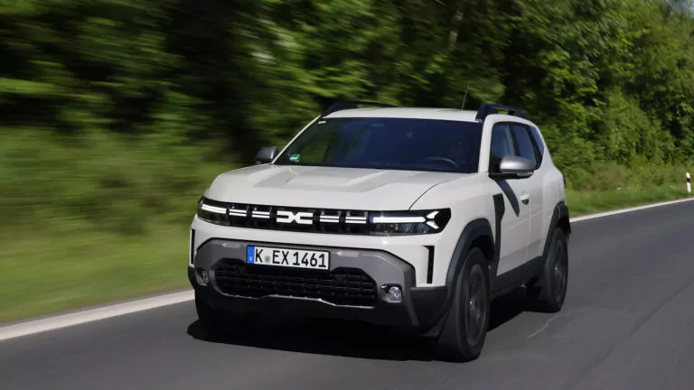 Ein weißer Geländewagen mit schwarzen Akzenten fährt auf einer glatten, von grünen Bäumen gesäumten Straße, wobei Bewegungsunschärfen seine Geschwindigkeit unterstreichen. Das Fahrzeug verfügt über einen modernen Frontgrill und LED-Scheinwerfer.