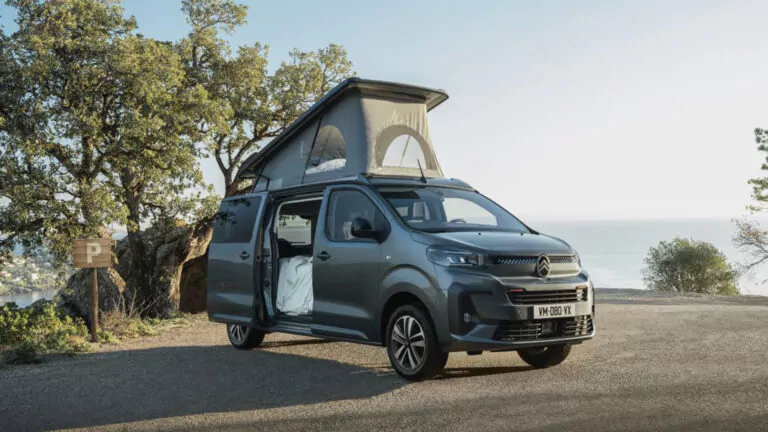 Ein graues Wohnmobil mit ausgefahrenem Aufstelldach und geöffneter Seitentür parkt an einer Küstenstraße, im Hintergrund sind Bäume und das Meer bei klarem Himmel zu sehen.