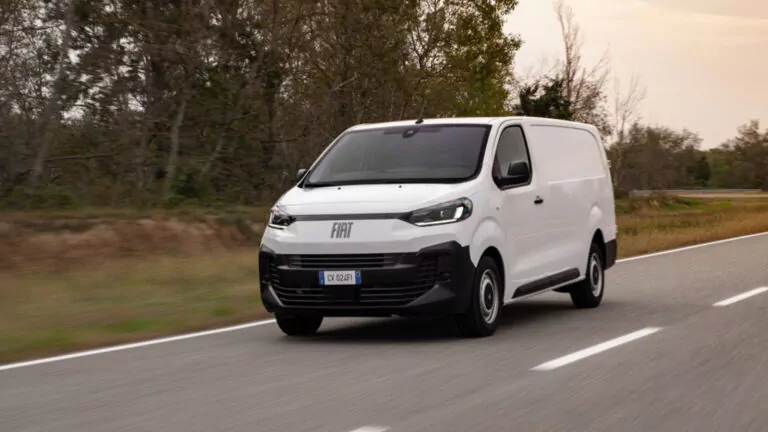 Ein weißer Fiat-Kastenwagen fährt auf einer von Bäumen und Gras gesäumten Landstraße unter einem bedeckten Himmel.