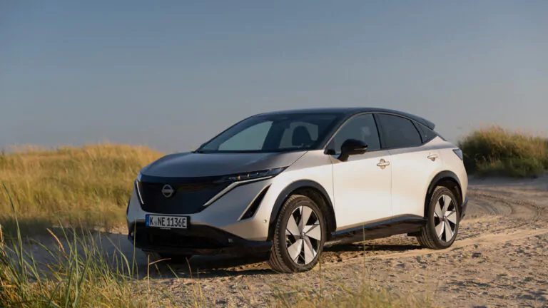 Ein weiß-schwarzer Nissan Ariya Elektro-Geländewagen, geparkt auf sandigem Gelände mit hohem Gras und einem klaren Himmel im Hintergrund.