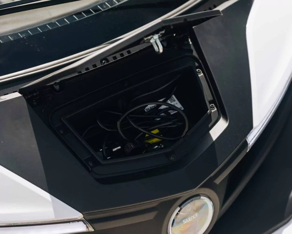 Nahaufnahme eines offenen Fachs an der Vorderseite eines Nissan Silence Angebot-Fahrzeugs, in dem aufgerollte Ladekabel und Stecker zu sehen sind. Das Äußere des Fahrzeugs ist in einem eleganten schwarz-weißen Design gehalten.