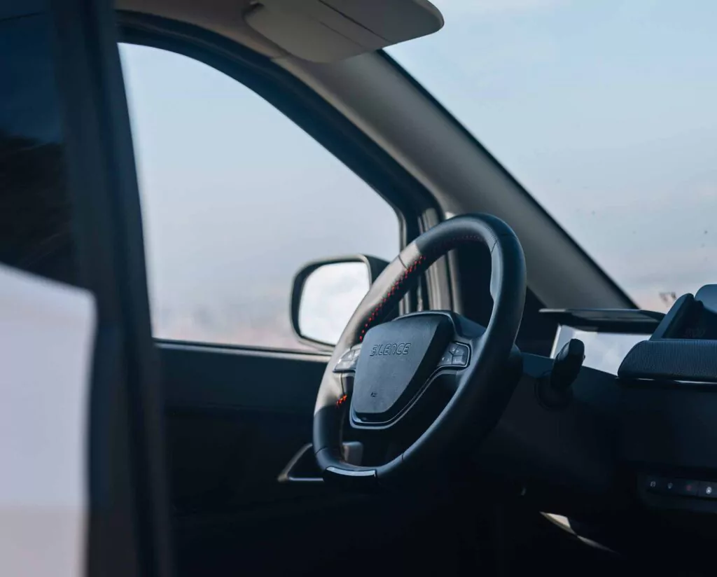 Nahaufnahme des Lenkrads und des Armaturenbretts eines Nissan Silence Angebot, mit geöffneter Fahrertür. Der Seitenspiegel und ein Teil des Fahrzeugs sind vor einem unscharfen Außenhintergrund zu sehen.