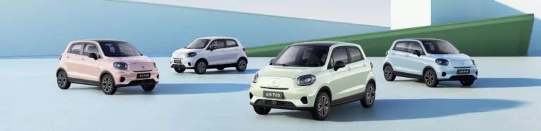 Fünf kompakte Elektroautos in Pastellfarben (rosa, weiß, mintgrün, hellblau und silber) sind auf einer glatten, offenen Fläche mit modernen, minimalistischen Architekturelementen im Hintergrund geparkt.