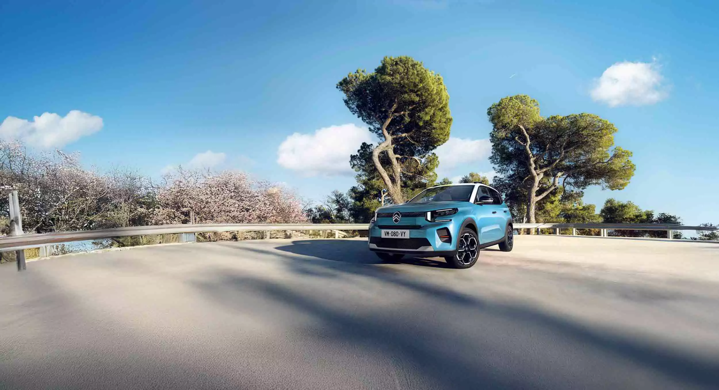 Ein blauer Renault Geländewagen parkt auf einer sonnenbeschienenen Betonstraße mit Metallgeländern, während ein Citroen C3 You vorbeifährt, umgeben von grünen Bäumen und einem blauen Himmel mit vereinzelten Wolken.