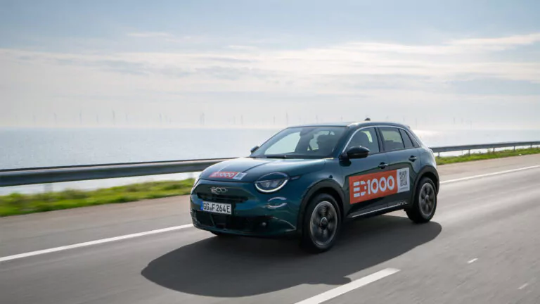 Ein dunkelgrünes Elektroauto mit "E|1000"-Schriftzug fährt auf einer Autobahn neben einem großen Gewässer, in der Ferne sind Windturbinen unter einem blauen Himmel mit vereinzelten Wolken zu sehen.