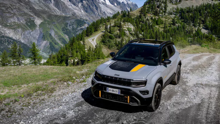 Ein grauer Jeep-Geländewagen mit schwarzen und gelben Details ist auf einer kurvenreichen, unbefestigten Bergstraße geparkt, umgeben von grünen Bäumen, grasbewachsenen Hügeln und felsigen Gipfeln im Hintergrund.