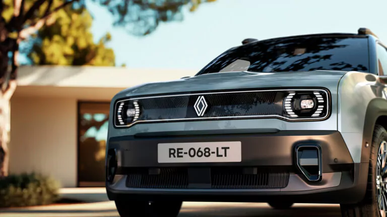 Nahaufnahme der Vorderseite eines modernen silbernen Renault, der im Freien geparkt ist, mit einem schnittigen Kühlergrill, deutlichen Scheinwerfern und einem sichtbaren Nummernschild mit der Aufschrift RE-068-LT. Ein Haus und Bäume sind unscharf im Hintergrund zu erkennen.