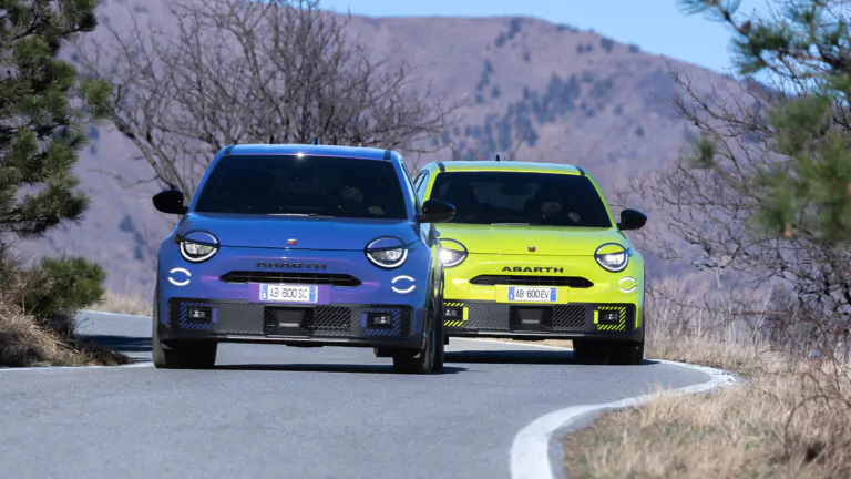 Zwei Abarth-Autos, ein blaues und ein gelbes, fahren Seite an Seite auf einer kurvenreichen Bergstraße mit trockenem Gras und Hügeln im Hintergrund.