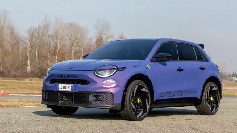 Ein lilafarbener Abarth Crossover-Geländewagen mit schwarzen Rädern, getönten Scheiben und einem auffälligen Abarth-Schriftzug an der Seite ist auf einer Straße geparkt. Im Hintergrund sind blattlose Bäume und ein bewölkter Himmel zu sehen.