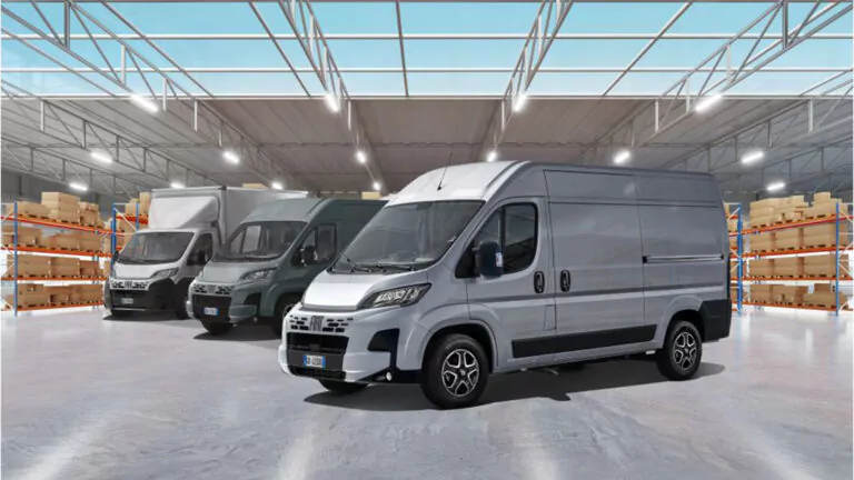 In einem geräumigen, gut beleuchteten Lagerhaus mit hohen Decken und mit Kisten bestückten Regalen im Hintergrund sind drei Lastwagen geparkt.