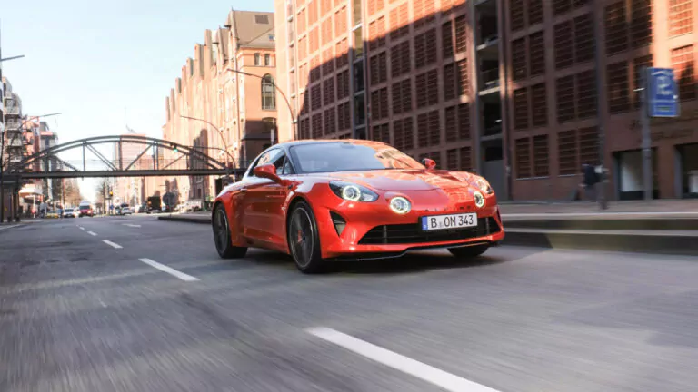 Ein leuchtend orangefarbener Sportwagen fährt an einem sonnigen Tag schnell eine von modernen Backsteingebäuden gesäumte Straße entlang, über die eine Fußgängerbrücke aus Metall führt.