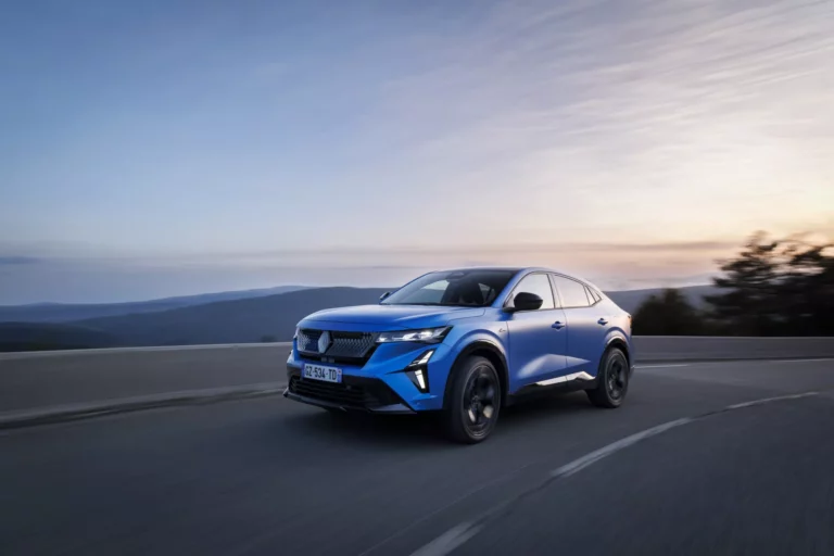 Ein blauer Geländewagen mit Nextory im Renault fährt bei Sonnenuntergang eine kurvige Bergstraße entlang, wobei seine unscharfe Bewegung die Geschwindigkeit betont. Der Himmel ist klar mit weichen Wolken, und die entfernten Hügel bieten eine atemberaubende Kulisse.