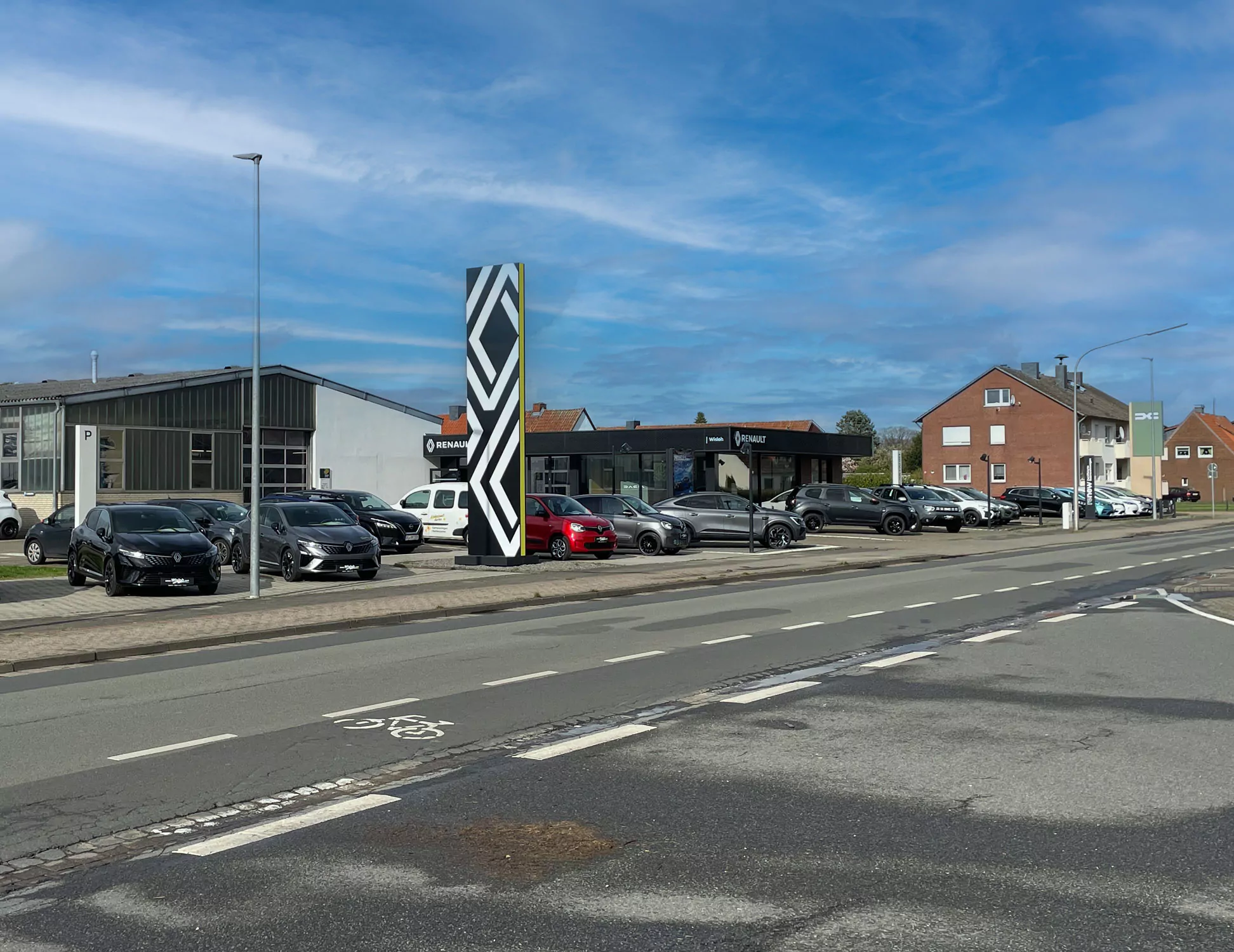 Ein Autohaus mit mehreren geparkten Autos im Freien, einem großen vertikalen Schild mit einem geometrischen Logo und modernen Gebäuden im Hintergrund unter einem teilweise bewölkten blauen Himmel.