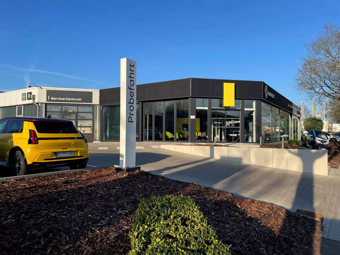 Ein modernes Autohaus mit großen Glasfenstern und schwarz-gelben Akzenten. Ein gelbes Auto ist draußen geparkt. Neben dem Eingang steht ein Schild mit der Aufschrift "Probefahrt". Unter dem blauen Himmel sind Bäume und Grünanlagen zu sehen.
