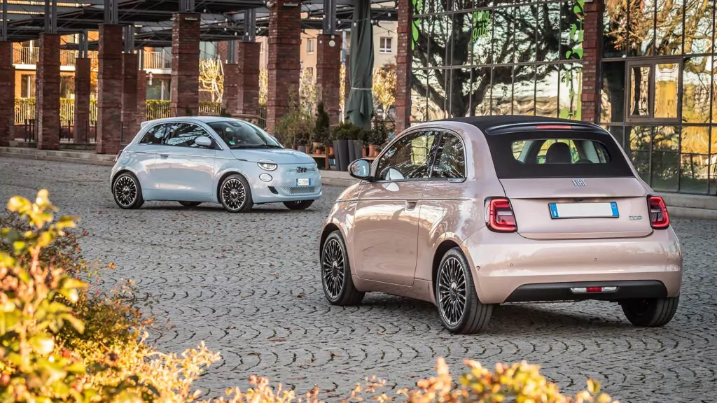 Zwei kleine, moderne Fiat-Modelle, darunter der stilvolle Fiat 500e Elektroauto LaPrima, stehen auf einem Kopfsteinpflaster vor einem Gebäude mit großen Fenstern und Backsteinsäulen. Im Hintergrund sind Bäume und Sträucher zu sehen.