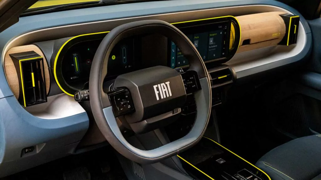 Nahaufnahme des Innenraums eines Fiat Grande Panda mit modernem Armaturenbrett, digitalen Anzeigen, Holzakzenten und gelben Zierleisten sowie einem großen, zentralen FIAT-Logo auf dem Lenkrad.