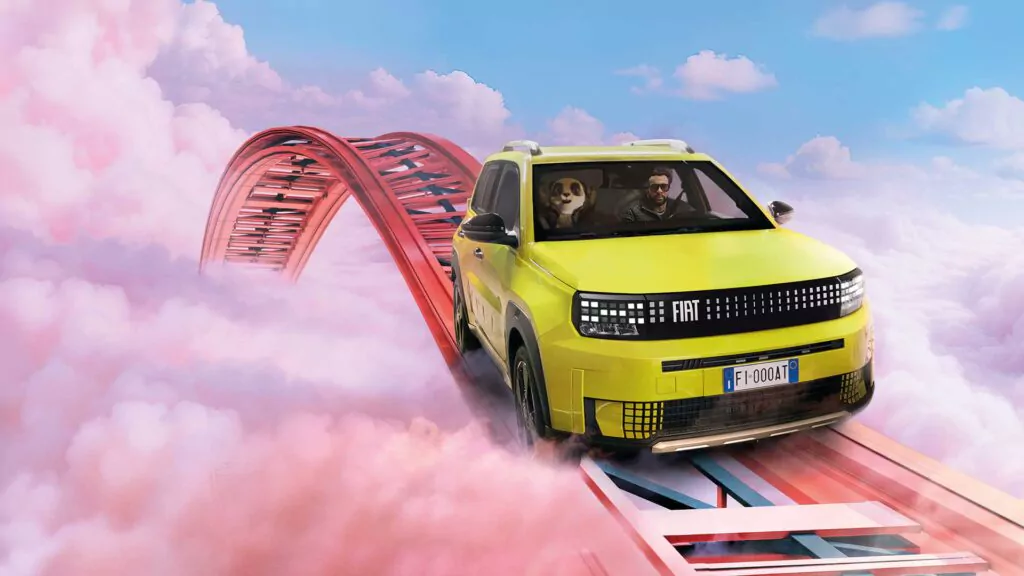 Ein gelber Fiat Grande Panda mit einem Mann und einem Hund darin fährt auf einer kurvenreichen Achterbahnstrecke hoch über rosa Wolken unter einem blauen Himmel.