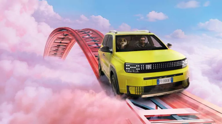 Ein gelber Fiat Grande Panda mit einem Mann und einem Hund darin fährt auf einer kurvenreichen Achterbahnstrecke hoch über rosa Wolken unter einem blauen Himmel.