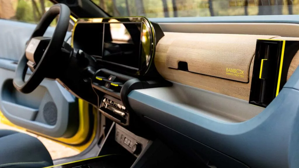 Nahaufnahme des modernen Innenraums eines Fiat Grande Panda mit digitalem Armaturenbrett, blauen und beigen Polstern und einer hölzernen Akzentplatte mit dem gelb hervorgehobenen Wort BAMBOO neben einem eingebauten Staufach.