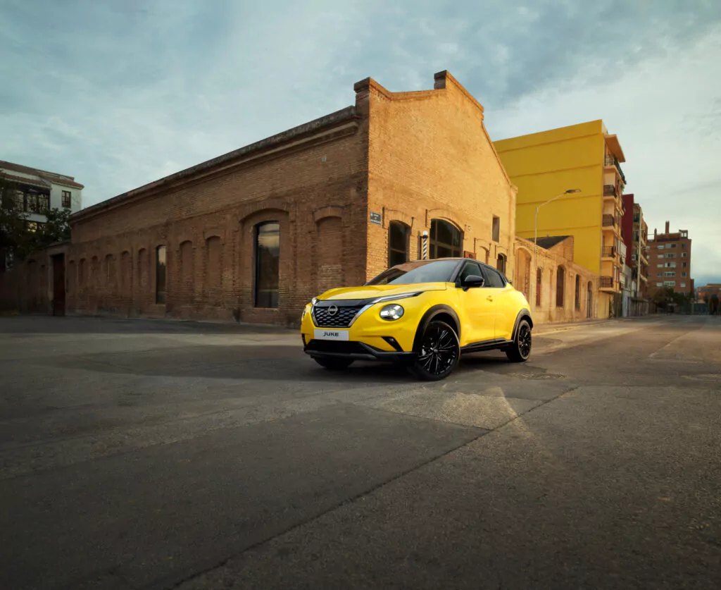 Ein leuchtend gelber Nissan JUKE N-CONNECTA mit schwarzen Rädern parkt auf einer ruhigen, leeren Straße vor einem alten Backsteingebäude unter einem wolkenverhangenen Himmel und hebt die neuesten Angebote in plakativem Stil hervor.