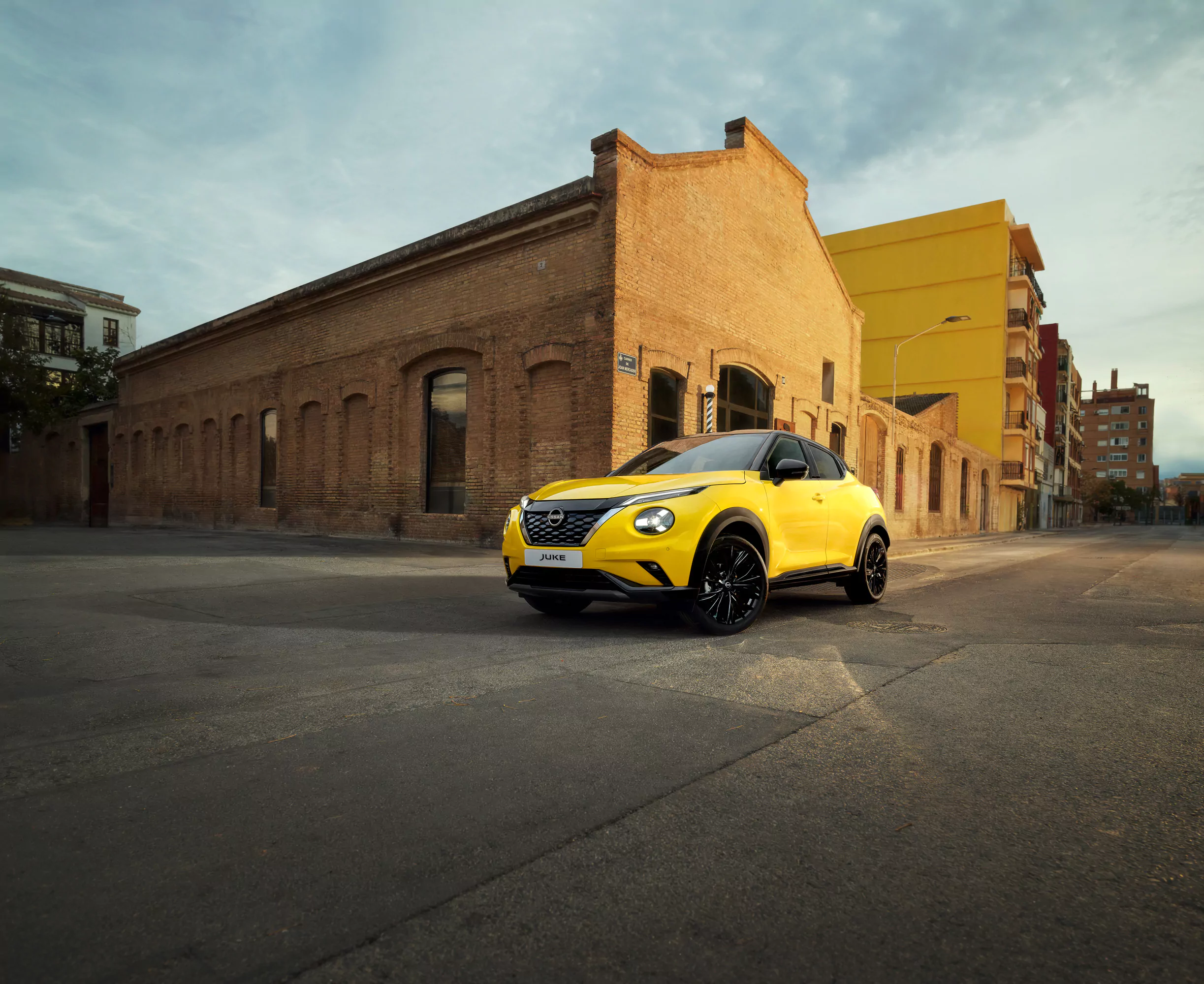 Ein leuchtend gelber Nissan JUKE N-CONNECTA mit schwarzen Rädern parkt auf einer ruhigen, leeren Straße vor einem alten Backsteingebäude unter einem wolkenverhangenen Himmel und hebt die neuesten Angebote in plakativem Stil hervor.