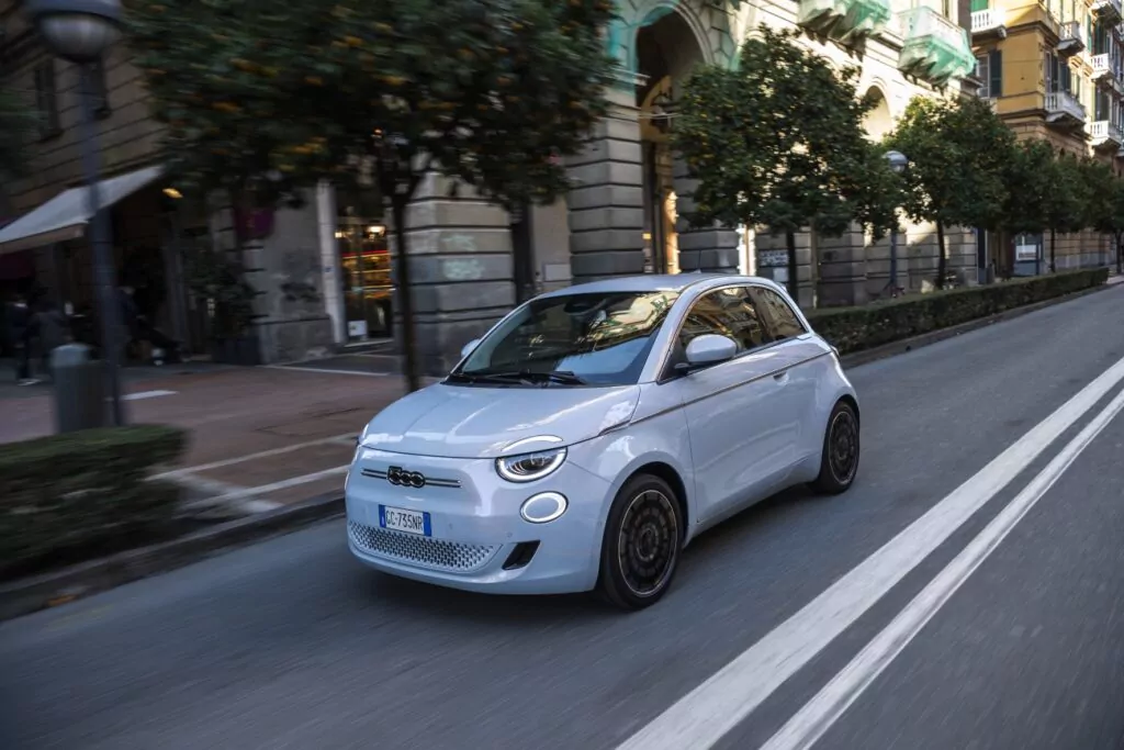Ein kleiner weißer 500e City Edition fährt eine von Bäumen und historischen Gebäuden gesäumte Straße entlang, im Hintergrund sind Geschäfte und Balkone zu sehen.