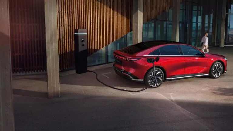 Ein rotes Mazda 6e-Elektroauto ist an einer Ladestation in der Nähe eines modernen Gebäudes mit großen Glasfenstern und Holzpaneelen geparkt. Das Auto ist eingesteckt und wird aufgeladen, während im Hintergrund eine Person spazieren geht.