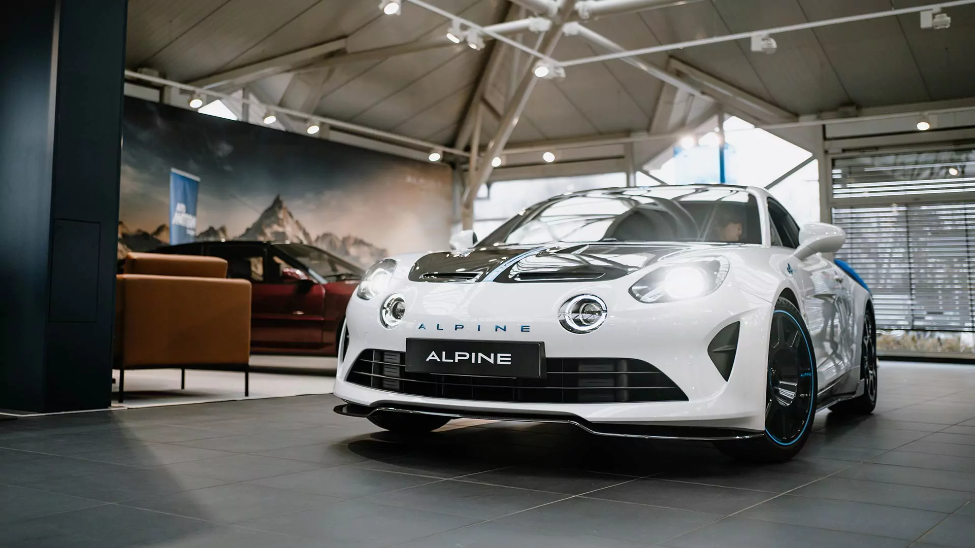 Ein weißer Alpine-Sportwagen wird in einem Ausstellungsraum ausgestellt, mit moderner Beleuchtung, großen Fenstern und einem anderen Auto, das teilweise im Hintergrund zu sehen ist.