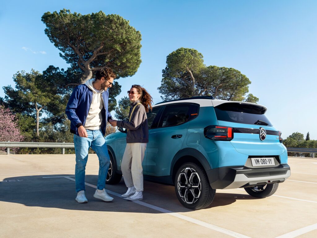 Ein lächelnder Mann und eine lächelnde Frau stehen zusammen neben einem geparkten hellblauen Citroen C3 You auf einem Parkplatz im Freien, mit Bäumen und einem klaren blauen Himmel im Hintergrund.