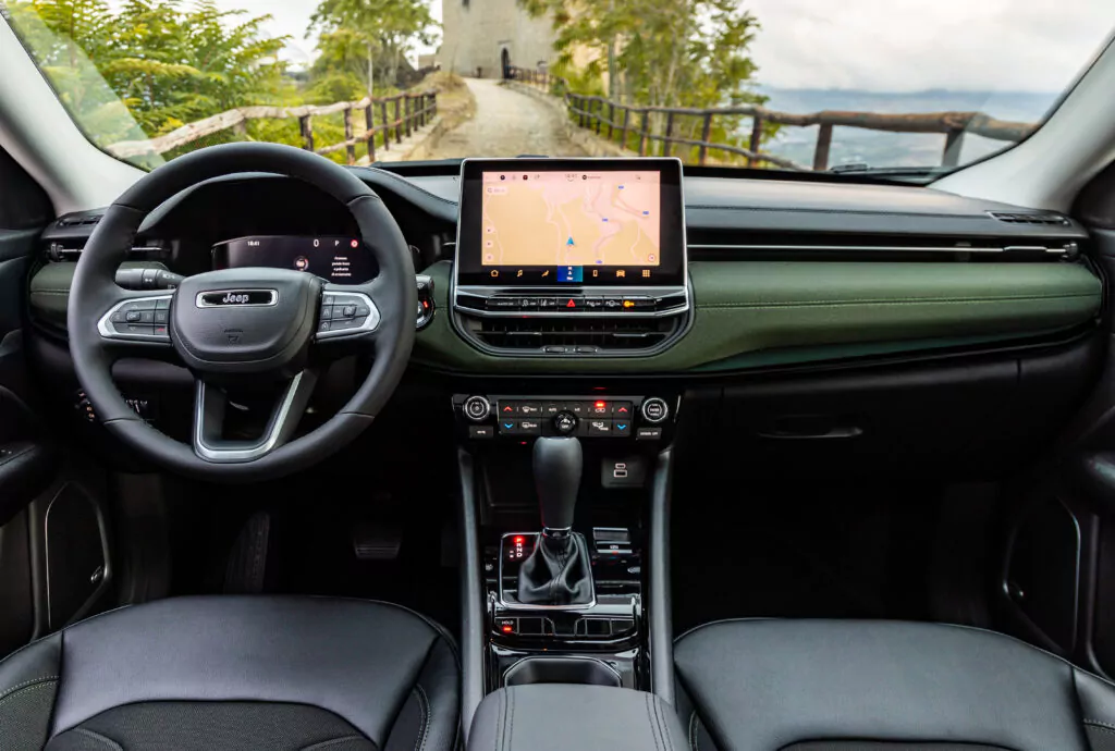 Ansicht des Innenraums eines Jeep-Fahrzeugs mit Lenkrad, Armaturenbrett, Touchscreen-Display mit Navigation, Schalthebel und Vordersitzen, wobei durch die Windschutzscheibe eine malerische Straße im Freien zu sehen ist.